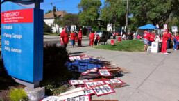 Nurses in red stand on picket line in background with hospital sign in foreground