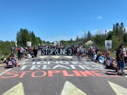 An outdoor Stop Line 3 protest with chalking in the foreground and a banner in the background
