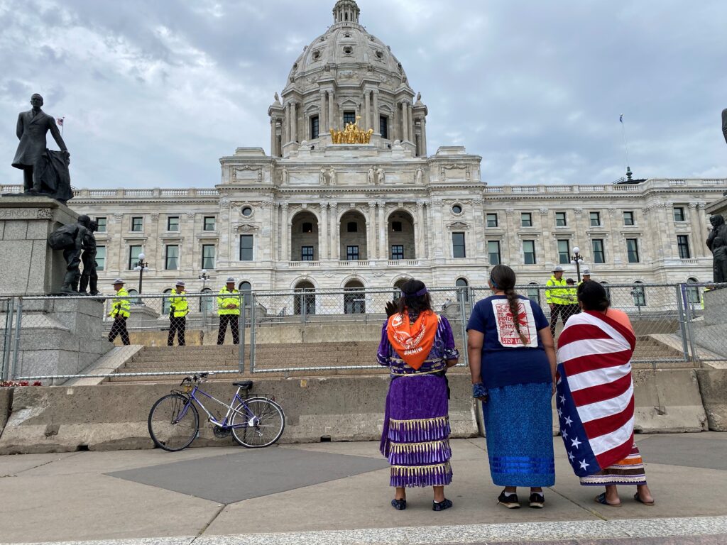 indigenous water protectors stand in front of a fenced off capitol filled with Police