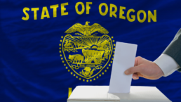 state of Oregon flag behind hand dropping ballot into voting box