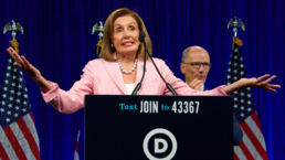 Nancy Pelosi shrugs at a podium with Tom Perez and American flags in the background