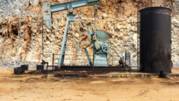 A rusted oil derrick sits next to a large barrel against a rock background showing layers of sediment
