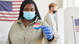 Blurred portrait of African-American woman holding I VOTED sticker while standing t polling station on post-pandemic election day