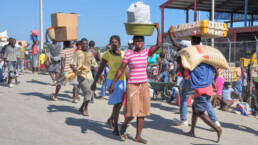 Haitian woman carries a bucket of goods on her head while men carry a poultry crate and sack of grains at Haitian Dominican Republic border market.