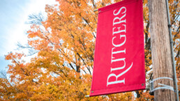 Rutgers University logo flag with backdrop of colorful maple tree leaves turning gold and red in autumn.