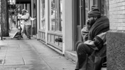 A homeless man perches on the street while men work in the background.