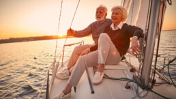 Beautiful and happy senior couple in love sitting on the side of sailboat or yacht deck floating in sea at sunset and enjoying amazing view, sailing together