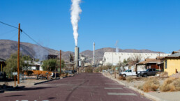 A view down a residential street in Trona with a prominent smokestack releasing emissions into the air.