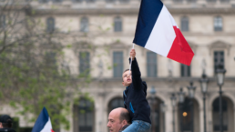 a small boy sits on his father's shoulders holding the french flag